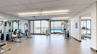 Located at 3033 W 71st Ave in Westminster, Colorado, this fitness facility spans a single floor and is thoughtfully divided into two dedicated exercise rooms, offering a comprehensive workout environment. Both rooms feature light wood-look flooring and crisp white walls accented by a drop-tile ceiling with flush-mounted linear fluorescent lighting that ensures bright, even illumination throughout.

The primary exercise room is highlighted by expansive floor-to-ceiling windows along one wall, flooding the space with natural light and providing sweeping views of the surrounding Westminster area, including a vivid blue body of water visible in the distance. This room houses a variety of strength-training equipment, including a multi-function cable machine, weight bench, and free-weight rack with dumbbells, along with a full-length mirror wall that enhances the sense of openness.

The secondary exercise room is equally spacious and features large windows along with glass-panel double doors that connect to the adjacent area. It is equipped with cardio machines, including treadmills and an elliptical, as well as additional strength equipment and balance tools such as a BOSU ball. The mirrored wall in this room further amplifies the light and generous sense of space. Together, the two rooms create a cohesive, modern fitness environment suited for a wide range of exercise routines.