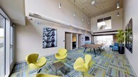 This contemporary space combines striking interior design with impressive outdoor amenities across a single thoughtfully designed floor. The interior lounge commands attention with its soaring ceilings fitted with exposed industrial-style ductwork and a series of pendant lights that illuminate the space from above. A ceiling fan adds to the airy atmosphere, while large windows flood the room with natural light. The lounge is furnished with a bold collection of chartreuse swan-style chairs arranged around a sleek metallic side table, set atop a vibrant patterned carpet that anchors the seating area in color and texture. A large black-and-white photographic artwork adorns one wall, adding an urban artistic element. A wooden conference or gathering table sits toward the rear, alongside access to additional rooms and corridors. The rooftop terrace offers an expansive open-air retreat featuring a modern pergola structure with a louvered roof, a built-in bar counter with dark wood paneling, and multiple seating areas. Concrete pavers cover the terrace floor, and the surrounding urban skyline provides a dynamic backdrop. The space is well-equipped with amenity lighting and waste receptacles, making it suitable for both casual gatherings and organized events.