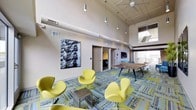 This contemporary space combines striking interior design with impressive outdoor amenities across a single thoughtfully designed floor. The interior lounge commands attention with its soaring ceilings fitted with exposed industrial-style ductwork and a series of pendant lights that illuminate the space from above. A ceiling fan adds to the airy atmosphere, while large windows flood the room with natural light. The lounge is furnished with a bold collection of chartreuse swan-style chairs arranged around a sleek metallic side table, set atop a vibrant patterned carpet that anchors the seating area in color and texture. A large black-and-white photographic artwork adorns one wall, adding an urban artistic element. A wooden conference or gathering table sits toward the rear, alongside access to additional rooms and corridors. The rooftop terrace offers an expansive open-air retreat featuring a modern pergola structure with a louvered roof, a built-in bar counter with dark wood paneling, and multiple seating areas. Concrete pavers cover the terrace floor, and the surrounding urban skyline provides a dynamic backdrop. The space is well-equipped with amenity lighting and waste receptacles, making it suitable for both casual gatherings and organized events.