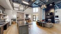 Located at 2811 S Cooper St in Arlington, Texas, this sophisticated single-floor clubhouse space showcases a stunning blend of modern and transitional design. Soaring double-height ceilings define the main gathering areas, accentuated by bold coffered detailing finished in deep navy and black, complemented by statement pendant and chandelier lighting. Wide-plank wood-look flooring flows continuously throughout, unifying the various zones of the space. A dramatic floor-to-ceiling dark metal fireplace column serves as the centerpiece of the main lounge, which is furnished with curated seating arrangements and anchored by a large-screen television. Oversized windows and glass doors flood the interior with natural light and offer views of the surrounding outdoor courtyard. A stylish wet bar area features dark cabinetry with brass hardware, granite countertops, open shelving, and a textured tile backsplash. Steel-framed glass partitions elegantly divide additional seating and lounge zones, including areas appointed with patterned wallcoverings and intimate booth-style seating. A substantial granite-topped island anchors the central gathering space. The overall ambiance is refined yet welcoming, balancing bold architectural moments with warm, carefully curated furnishings throughout.