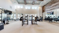 This fitness center spans a generous single floor, thoughtfully divided into two functional workout zones. The main exercise area commands attention with its dramatic double-height ceilings accented by coffered detailing and exposed wood beam inserts. A striking reclaimed wood accent wall adds warmth and texture, contrasting beautifully with the crisp white walls. Clerestory windows positioned along the upper walls flood the space with natural light, while large floor-level windows offer outdoor views and enhance the bright, open atmosphere. Full-length mirrors line portions of the walls, amplifying the sense of space. This primary area is equipped with a comprehensive array of strength and cardio machines, including treadmills, stair climbers, cable machines, weight benches, and a dumbbell rack. A separate studio room, finished in soft blue-grey tones, provides a dedicated space for cycling and group fitness activities. Fitted with True cycling bikes, a wall-mounted TV, stability balls, medicine balls, kettlebells, and yoga mats, this room also features ceiling fans and its own mirror wall. Neutral carpeted flooring throughout both areas contributes to a clean, cohesive aesthetic across the entire facility.
