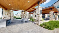 This expansive outdoor amenity space is defined by its striking architectural design, combining natural limestone columns with a warm, tongue-and-groove wood ceiling supported by exposed timber beams. Recessed lighting and ceiling fans are integrated throughout the covered pavilion, ensuring comfort across all seasons. The generous floor area transitions between light hardwood-style tile underfoot in the lounge zones and classic paver stone throughout the open-air sections, lending visual variety and texture to the space. Multiple distinct seating areas accommodate a range of social activities — vibrant green Adirondack chairs cluster around a casual lounge area, while wicker sectional sofas and coffee tables create a more relaxed gathering zone. Dining tables with modern metal chairs provide a dedicated space for outdoor meals. Substantial stone columns frame open sightlines to the surrounding landscaping, iron fencing, and the resort-style pool deck beyond, where lounge chairs and umbrellas line the water's edge. Lush plantings of ornamental grasses and shrubs soften the hardscape throughout. The pavilion connects seamlessly to adjacent outdoor areas and is framed by views of the surrounding multi-story residential buildings, creating a cohesive, community-centered environment ideal for both lively gatherings and quiet relaxation.