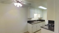 This single-floor space offers a clean and functional layout centered around a well-appointed kitchen. The kitchen features classic white cabinetry — both upper and lower — providing ample storage for everyday essentials. Dark stone-style countertops create a striking contrast against the white cabinets, adding a touch of visual depth to the space. A double-basin stainless steel sink sits beneath a convenient under-cabinet area, keeping the workspace organized and efficient. A recessed skylight panel in the ceiling floods the kitchen with bright, even overhead lighting, enhancing the open and airy feel of the room. A ceiling fan with a light fixture is positioned nearby, ensuring comfortable air circulation throughout. The light-toned tile flooring and neutral wall tones contribute to a cohesive, understated aesthetic that makes the space feel clean and easy to personalize. The overall design is straightforward and practical, offering a solid foundation for comfortable day-to-day living.