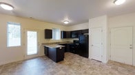 This single-story space showcases a well-appointed kitchen as its central feature. The kitchen is designed with dark espresso-toned cabinetry that extends to the upper and lower walls, creating a bold contrast against the light, neutral-colored walls. A peninsula configuration adds extra counter space and enhances the kitchen's functionality, making it suitable for both meal preparation and casual use.

The kitchen is equipped with a full suite of black appliances, including a refrigerator, electric range, and an over-the-range microwave, providing a cohesive and modern aesthetic. The countertops feature a speckled granite-style finish that complements the dark cabinetry. A sink is positioned beneath a window fitted with horizontal blinds, allowing natural light to filter into the space.

The flooring throughout is a durable tile in a warm, neutral tone, adding to the overall practicality of the space. A panel door leads to an adjacent room, suggesting additional living areas beyond the kitchen. Two exterior-facing windows and a glass-paneled door contribute natural light, maintaining a bright and open atmosphere despite the deep-toned cabinetry.