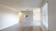 This well-appointed single-story, one-bedroom, one-bathroom unit offers a practical and comfortable layout across a thoughtfully organized floor plan. The combined kitchen and living area forms the heart of the home, featuring light-toned luxury vinyl plank flooring, neutral gray walls, and a textured ceiling throughout. A charming pass-through window connects the kitchen to the living room, enhancing the open feel of the space. The galley-style kitchen is equipped with white cabinetry featuring brushed nickel hardware, light countertops, a stainless steel sink, a dishwasher, and black appliances including an electric range and refrigerator, all illuminated by overhead lighting. Natural light filters into the living area through window blinds, adding brightness to the neutral palette. The bedroom is generously sized, finished with plush carpet, a ceiling fan with light kit, and a window for natural light. A walk-in closet with built-in shelving and hanging rods provides ample storage. The full bathroom features a tub-shower combo with white tile surround, wood-look vinyl flooring, a toilet, and a vanity with a marble-style countertop. Clean finishes and a cohesive neutral color scheme give the entire unit a fresh, move-in-ready appearance.