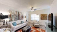 This single-floor space centers around a well-appointed living room designed for both comfort and style. The room is finished with light-colored walls and a smooth white ceiling, from which a classic five-blade ceiling fan with integrated lighting is mounted, providing both air circulation and ambient illumination. A generously sized window fitted with horizontal blinds allows natural light to fill the room, creating a bright and airy atmosphere.

The living area is furnished with a crisp white sofa accented by teal and patterned throw pillows, complemented by a coordinating accent chair and ottoman in a neutral print fabric. A bold burnt-orange area rug with an ornate scroll motif anchors the seating arrangement, adding a vibrant focal point to the space. A pair of large-format abstract artworks adorns one wall, contributing color and visual interest, while an arc floor lamp provides additional task lighting near the seating area.

The flooring appears to be neutral-toned carpet, adding warmth underfoot throughout the room. Overall, the space presents a clean, contemporary aesthetic with thoughtful decorative touches that create an inviting and livable environment.