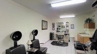 This single-floor hair salon offers a clean, functional environment designed to accommodate a range of hair care services. The space is finished with light-toned walls and white tile flooring, creating a bright and hygienic atmosphere complemented by overhead fluorescent lighting that ensures consistent illumination throughout.

The styling area is equipped with a classic hydraulic stylist's chair positioned in front of a large wall-mounted mirror, providing ample workspace for the stylist. A well-organized station alongside the mirror holds professional hair care products and tools, keeping essentials within easy reach. A rolling utility cart nearby adds additional storage and flexibility for equipment and supplies.

On the opposite side of the room, a hooded hair dryer station features a cushioned client chair and a small side table, creating a comfortable waiting and treatment area. Framed artwork on the walls adds a personal touch, contributing to a welcoming ambiance. A laundry hamper is also visible, reflecting the practical, day-to-day operational needs of an active salon. The overall layout is efficient and well-organized, making effective use of the available space while maintaining a tidy and professional environment suitable for both stylists and clients.