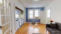 Located at 4 E 32nd Street in Baltimore's vibrant neighborhood, this single-floor unit offers a functional and characterful living space. The main living area is highlighted by warm, honey-toned hardwood floors that run throughout, crisp white trim, and a bold blue accent wall that adds a pop of color and personality. A ceiling fan overhead provides comfort, while a double-hung window allows natural light to fill the room. The open layout connects the living space to a compact, efficiently designed kitchen tucked into an alcove. The kitchen features gray upper cabinetry, white appliances including a gas range and refrigerator, and under-cabinet lighting. The bathroom retains classic charm with white subway tile wainscoting, a mosaic tile floor in a retro checkerboard and mosaic pattern, a wall-mounted porcelain sink, and a medicine cabinet mirror. The living room provides a comfortable and versatile space with clean white walls, additional natural light from a side window, and ample room for seating and storage. Throughout the unit, the consistent hardwood flooring, neutral palette, and vintage tile details come together to create a cohesive and practical living environment well-suited for everyday life in Baltimore.