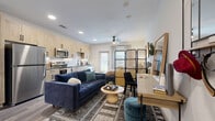This thoughtfully designed studio unit offers an efficient single-floor layout that maximizes every square foot through smart spatial planning and modern finishes. The open-concept main area seamlessly integrates the kitchen, living, and sleeping zones into one cohesive space. A rattan-panel room divider provides a stylish visual separation between the sleeping area and the living room, which features wood-look plank flooring, recessed lighting, and a ceiling fan. The living area is anchored by a patterned area rug and opens toward a glass door that introduces natural light from outside. A built-in wall-mounted desk with a mirror serves as a dedicated workspace near the entryway, which also features a wall-mounted hook rack for added convenience. The kitchen is fully equipped with stainless steel appliances — including a refrigerator, electric range, over-the-range microwave, and dishwasher — complemented by light wood cabinetry with matte black hardware, a white subway tile backsplash, and dark countertops. The bathroom offers a full tub-shower combination, wood-toned vanity cabinetry with dark countertops and matte black fixtures, and tile flooring. A closet completes the layout, providing dedicated storage within this compact yet well-appointed space.