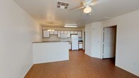 This single-floor unit features an open-concept design that seamlessly connects the kitchen and living area into one cohesive space. The kitchen is outfitted with white cabinetry accented by warm wood-toned trim, offering generous storage across upper and lower cabinets. A white refrigerator, microwave, and range provide essential appliances, while a peninsula countertop with a dark stone-look surface serves as a natural divider between the kitchen and the main living area. A pendant chandelier hangs above the peninsula, adding a decorative touch to the kitchen zone. The living area benefits from a ceiling fan with a light fixture, contributing to both comfort and ambient lighting. Throughout the open space, warm wood-look laminate flooring creates a cohesive, easy-to-maintain surface. The walls are finished in a clean white, keeping the interior feeling bright and open. A doorway visible from the main area leads to additional rooms within the unit. The overall layout is practical and functional, maximizing the use of the shared living and kitchen space within a compact, single-level footprint.