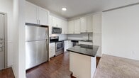Unit 429 at 4699 Kittredge Street in Denver, Colorado offers a well-designed 660-square-foot, single-floor layout comprising one bedroom, one bathroom, a combined kitchen and living room, a hallway, and a closet. The kitchen is a standout feature, outfitted with stainless steel appliances including a refrigerator, electric range, and over-the-range microwave. White shaker cabinetry, a subway tile backsplash, dark granite countertops, and a central island with breakfast bar seating complement the rich hardwood-style flooring throughout the kitchen area. The open living room extends from the kitchen and is finished with plush carpet and crown molding trim, offering a bright, neutral palette. French doors lead to a private balcony, filling the living space with natural light and outdoor views. The 150-square-foot bedroom features carpet flooring, a large window with exterior views, and a closet with double panel doors. The bathroom includes a dark granite-topped vanity with white cabinetry, gray subway tile wainscoting, a large mirror, and hardwood-style flooring, creating a clean and modern aesthetic. The unit's light-toned walls and consistent finishes throughout contribute to a cohesive and functional living environment.