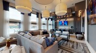 This sophisticated single-floor communal lounge is designed for comfort and social interaction, combining modern aesthetics with thoughtfully curated furnishings. The space is anchored by soaring ceilings adorned with oversized drum pendant lights that cast a warm, inviting glow throughout. Expansive floor-to-ceiling windows dressed with layered sheer and navy drape panels flood the interior with natural light while offering views of an outdoor pool area.

The main lounge area features generously sized sectional sofas and accent chairs arranged around a herringbone-top coffee table, all set atop a richly patterned area rug. A billiards table is positioned near the windowed wall, serving as both a functional and stylish focal point. Multiple distinct seating vignettes are distributed throughout, including a quieter lounge corner with a wall-mounted television, sleek accent chairs, and a dining-height table with upholstered seats.

The wet bar area is finished with dark cabinetry, matte countertops, brass hardware, a stainless steel sink, and a decorative patterned tile backsplash — all illuminated by industrial-style pendant lights. The overall palette of warm grays, navy blues, and gold accents lends the space a refined, contemporary ambiance suited for both relaxation and entertaining.