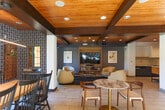 Located at 200 Winchester Cir in Los Gatos, CA, this thoughtfully designed single-floor space seamlessly integrates a living room, dining room, and kitchen into a cohesive, open layout. Striking wood-paneled ceilings with exposed dark wood beams lend warmth and architectural character throughout, complemented by recessed lighting and light tile flooring.

The living area is elegantly furnished with cream leather lounge chairs and a curved sofa arranged around a geometric gold-framed glass coffee table. A built-in entertainment center with shelving anchors the space, while bold patterned wallpaper adds a distinctive design accent. Large black-framed glass doors open directly to an outdoor courtyard, flooding the room with natural light and views of lush landscaping.

The dining area features a generous table surrounded by classic Windsor-style chairs beneath the same warm wood-beamed ceiling. A large window frames views of an outdoor fountain, creating a bright and inviting atmosphere.

The kitchen is outfitted with sleek, dark lower cabinetry, quartz countertops with a marble-look backsplash, stainless steel appliances including a range, dishwasher, and refrigerator, and a central island providing ample workspace. A warm wood open shelf adds a finishing touch above the counters. The overall ambiance is modern, welcoming, and well-appointed.