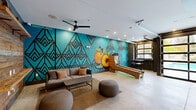 Located at 960 Marietta St NW in Atlanta's Westside neighborhood, this dynamic amenity lounge is a standout communal space defined by bold design and local character. The room's most striking feature is a sweeping floor-to-ceiling mural by a local artist, rendered in vivid teal with geometric Art Deco-inspired diamond patterns and a signature Georgia peach motif — a clear nod to Atlanta's identity. Polished concrete floors run throughout, lending an industrial yet clean aesthetic that complements the space's modern edge. A skee-ball lane anchors the entertainment zone, accompanied by high-top bar tables and stools for casual socializing. A comfortable lounge area features a neutral sofa, woven area rug, and rounded ottoman poufs, creating an inviting space for relaxation. Recessed ceiling lighting and a ceiling fan ensure the room remains well-lit and comfortable. On the opposite end, a built-in wet bar with open shelving and a mini-refrigerator provides convenience for gatherings, flanked by a reclaimed wood accent wall bearing a stylized "Atlanta" marquee sign. Full-panel glass garage doors open entirely to reveal a pool area beyond, flooding the space with natural light and creating a seamless indoor-outdoor connection. A teal accent lounge chair adds a pop of color near the entrance, tying the entire palette together cohesively.