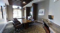 This professional conference space combines refined design with functional elegance. The centerpiece of the room is a stunning live-edge wooden conference table, which anchors the space and seats a generous number of attendees in comfortable upholstered rolling chairs. The ceiling draws the eye upward with bold exposed dark wood beam detailing, complemented by stylish industrial-style pendant lights that add warmth and character to the room.

Natural light pours in through large windows dressed with sheer curtains, creating a bright and welcoming atmosphere while offering views of the outdoors. The walls are finished in a rich, deep brown and neutral gray palette, lending the space a polished, corporate aesthetic. A wall-mounted flat-screen display serves as a focal point for presentations, while a credenza provides additional storage and surface space for meetings.

The flooring features a patterned carpet in earthy tones, adding texture and acoustic comfort underfoot. Framed artwork on the walls contributes a curated, professional touch to the overall ambiance. The thoughtful combination of natural materials, modern technology, and sophisticated design details makes this conference room a highly functional and visually impressive environment for business gatherings of all kinds.