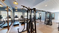Located on Legends Drive in Southern Pines, NC, this well-appointed exercise facility offers a generously sized, single-floor layout designed to accommodate a full range of fitness activities. The space is divided into distinct functional zones, seamlessly connected to create an open yet organized environment.

The strength and cardio area features durable, neutral-toned flooring and is equipped with a comprehensive selection of machines, including treadmills, ellipticals, cable systems, a power rack, and weight benches. Soft blue walls, recessed lighting, and large windows allow natural light to filter in, creating an energizing atmosphere with views of the surrounding greenery.

A dedicated studio zone transitions to warm hardwood-style flooring and is accented by exposed white wood-beam ceilings with shiplap detailing, lending a refined, architectural character to the space. Full-length mirrors in rich wood frames line the walls, amplifying the sense of openness, while wall-mounted sconce lighting adds warmth. This area is well-suited for stretching, yoga, or group fitness activities.

Throughout the facility, white shiplap ceiling treatments and soft blue-gray walls maintain a cohesive, upscale aesthetic. French doors and interior windows connect zones fluidly, while organized storage solutions and a medicine ball rack ensure the space remains functional and tidy. The overall design balances practicality with polished, inviting finishes.