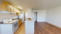 Located at 10589 14th Ave SW in Seattle, WA, this single-floor unit offers a functional and straightforward layout designed for comfortable everyday living. The main living area greets you at entry with warm wood-look flooring, a large window fitted with vertical blinds, and a ceiling light fixture, creating a bright and welcoming atmosphere with views of the surrounding greenery outside.

The kitchen is arranged in a galley style along one wall and opens to the living space via a raised peninsula counter — ideal for casual dining or meal prep. It features light maple cabinetry providing ample storage, a white electric range, a double-basin sink, and a full-size white refrigerator. Recessed under-cabinet fluorescent lighting illuminates the workspace, and a textured ceiling adds classic character throughout.

Adjacent to the kitchen, the living and sleeping areas are defined by continued wood-look flooring and clean white walls. The bedroom includes a built-in closet with shelving, offering practical storage. The bathroom is clean and simply appointed, with a full tub and shower combination, a toilet, a small frosted window for natural light, and durable tile-style flooring. The overall ambiance of the space is clean, neutral, and move-in ready.
