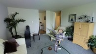 This single-floor residence is organized around an open living area finished with wall-to-wall carpet in a neutral grey tone, creating a soft, cohesive foundation throughout the space. The living area is furnished with a cushioned sofa accented by a patterned throw pillow, positioned near a glass-topped coffee table that holds decorative items including a vase of dried purple eucalyptus branches, adding a natural, organic touch to the room. Framed artwork on the walls contributes to a curated, lived-in aesthetic.

Adjacent to the living area, a compact dining nook features a round glass table paired with sleek black chairs, set on a hard-surface floor that distinguishes it from the carpeted main living zone. This transition between flooring materials subtly defines each functional area without the use of walls or dividers.

A structural column and open hallway are visible toward the back of the space, leading to additional rooms beyond. Potted plants and greenery are placed throughout, enhancing the interior with natural elements. The overall ambiance is calm and residential, with neutral wall tones and warm lighting contributing to an inviting atmosphere.