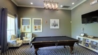 This single-floor space is designed as a refined recreational retreat, with the billiards room serving as its defining feature. At the heart of the room sits a full-size pool table with a rich dark wood finish and a black felt surface, resting atop a patterned geometric area rug that adds visual depth to the space. Overhead, an industrial-style chandelier with exposed Edison bulbs provides warm, focused lighting that enhances the room's sophisticated ambiance. The walls are finished in a cool, neutral gray tone and adorned with crown molding, lending an air of classic elegance. Decorative artwork, including a chandelier-inspired canvas print, complements the curated aesthetic. Two framed windows dressed with tailored Roman shades allow natural light to filter into the room, while a plush wingback armchair with a floral pattern offers a comfortable spot for spectators. Recessed ceiling lights provide additional illumination throughout. The overall atmosphere is one of polished leisure — a well-appointed space that balances functionality with tasteful interior design, making it an inviting environment for both recreation and relaxation.