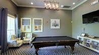 This single-floor space is designed as a refined recreational retreat, with the billiards room serving as its defining feature. At the heart of the room sits a full-size pool table with a rich dark wood finish and a black felt surface, resting atop a patterned geometric area rug that adds visual depth to the space. Overhead, an industrial-style chandelier with exposed Edison bulbs provides warm, focused lighting that enhances the room's sophisticated ambiance. The walls are finished in a cool, neutral gray tone and adorned with crown molding, lending an air of classic elegance. Decorative artwork, including a chandelier-inspired canvas print, complements the curated aesthetic. Two framed windows dressed with tailored Roman shades allow natural light to filter into the room, while a plush wingback armchair with a floral pattern offers a comfortable spot for spectators. Recessed ceiling lights provide additional illumination throughout. The overall atmosphere is one of polished leisure — a well-appointed space that balances functionality with tasteful interior design, making it an inviting environment for both recreation and relaxation.