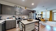 Located at 1700 W Frankford Rd in Carrollton, TX, this well-appointed one-bedroom, one-bathroom residence offers a thoughtfully designed single-floor layout. The open-concept kitchen and dining area is a standout feature, showcasing dark wood cabinetry extending to the ceiling, white quartz countertops, a tile backsplash, and a full suite of stainless steel appliances. A freestanding kitchen island provides additional prep and dining space. The kitchen flows seamlessly into the living room, which is bright and welcoming thanks to large windows with natural light and direct access to an outdoor area. Wood-style flooring runs throughout the main living spaces, adding warmth and continuity. The bedroom is a comfortable retreat featuring plush carpet, a ceiling fan, and generous windows. It connects directly to a hallway with two closets, including a large walk-in closet outfitted with multi-level shelving and hanging rods. The full bathroom is finished with dark cabinetry, a white vanity top, wood-style flooring, and a shower/tub combination. An in-unit laundry area adds everyday convenience. The overall design balances modern aesthetics with functional living throughout.