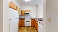 Located at 1200 Elm in Manchester, NH, this single-floor, one-bedroom, one-bathroom unit offers a practical and comfortable layout across a well-utilized footprint. The combined kitchen and living area forms the heart of the space, with the kitchen featuring warm-toned wooden cabinetry, a full suite of white appliances including a refrigerator, electric range, built-in microwave, and dishwasher, as well as a double-basin sink and ample counter space. Light wood-style flooring runs throughout the kitchen and living area, contributing to a bright, clean aesthetic. The living room is notably airy, bathed in natural light streaming through a large sliding glass door fitted with vertical blinds, which opens to an outdoor view and adds a sense of openness to the space. The bedroom is comfortably sized, finished with plush carpeting, and benefits from its own sliding glass door, also with vertical blinds, inviting additional natural light. A dedicated closet with a built-in rod provides convenient storage. The bathroom features wood-style flooring and is accessible directly from the main corridor. Neutral, warm-toned walls carry throughout the entire unit, creating a cohesive and welcoming ambiance.