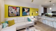 This well-appointed single-floor residence showcases a contemporary design aesthetic with a bold, energetic color palette and high-quality finishes throughout. The open-concept kitchen is a standout feature, boasting granite countertops, a two-tone cabinetry scheme combining white upper cabinets with wood-grain lower cabinets, stainless steel appliances including a refrigerator and range, and a peninsula with bar seating. Track lighting and pendant fixtures illuminate the space, while light wood-style flooring adds a clean, modern touch. A vibrant yellow accent wall connects the kitchen to the living area, where a white sectional sofa, bold abstract artwork, and layered lighting create an inviting atmosphere. The bedrooms are generously proportioned, featuring soft plush carpeting, ceiling fans, and ample natural light from large windows. One bedroom includes a walk-in closet, while decorative wall accents and mirrored elements add personality. The bathroom is finished with crisp white tile, a walk-in glass shower, granite vanity countertop, and wood-toned cabinetry, maintaining the cohesive modern style found throughout the residence. Overall, the space exudes a vibrant yet refined ambiance.