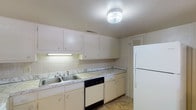 This single-floor, two-bedroom, one-bathroom unit offers a well-organized layout with a clear separation between living and sleeping areas. The kitchen is equipped with ample white cabinetry, a marble-patterned countertop, a double stainless steel sink, a dishwasher, a refrigerator, and a tile backsplash. A pass-through window connects the kitchen directly to the living area, creating an open, communicative flow between the two spaces.

The living area is a generous, carpeted room highlighted by a striking exposed brick accent wall that adds texture and character to the space. A sliding glass door floods the room with natural light and provides outdoor access, while a small chandelier adds a decorative touch. The kitchen and living areas share a cohesive, functional design.

Both bedrooms are carpeted and feature windows that allow natural light, creating a bright and comfortable atmosphere. The full bathroom includes tile flooring set in a diagonal pattern, white square wall tiles, a marble-patterned countertop vanity, and a tub/shower combination, presenting a clean, functional design throughout.