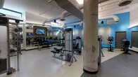 This fitness center combines industrial design with modern amenities to create a functional and visually dynamic workout space. The room features exposed ductwork and structural concrete columns that lend an industrial character, while bold blue accent walls add energy and visual interest throughout. The ceiling height provides an open, airy feel conducive to an active environment.

The space is well-appointed with a comprehensive selection of exercise equipment, including strength-training cable machines, a dedicated free weights area with a full dumbbell rack, and multiple cardio machines such as treadmills. A large flat-screen television is wall-mounted near the free weights section, offering entertainment during workouts. Floor-to-ceiling mirrors line one wall, enhancing the sense of space while allowing users to monitor their form.

The flooring is a smooth, light-colored surface suited for fitness use, and the lighting is bright and evenly distributed, ensuring good visibility across all areas. Textured mosaic-style wall panels add an additional design element. The overall layout is well-organized, providing clear zones for both strength training and cardio activity within a single, cohesive space.