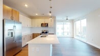 This contemporary single-floor residence showcases a thoughtfully designed open-concept layout with a cohesive modern aesthetic throughout. The kitchen serves as the centerpiece of the living area, featuring a large central island with a white quartz countertop and undermount sink, two-tone cabinetry pairing crisp white upper cabinets with warm wood-grain lower cabinets, and a full suite of stainless steel appliances including an electric range, over-the-range microwave, and French door refrigerator. Pendant lighting above the island adds a refined touch, while large windows in the adjacent living area flood the space with natural light and are paired with a ceiling fan for year-round comfort. Warm hardwood-style flooring runs consistently throughout the entire unit, creating a unified and inviting atmosphere. A well-lit hallway with recessed and flush-mount lighting connects the living areas to the private quarters. The bedrooms are generously proportioned, each equipped with ceiling fans, and several feature windows that frame striking urban skyline views. The bathrooms are finished with wood-toned vanity cabinetry, white quartz countertops, brushed nickel fixtures, and full shower-tub combinations, delivering a clean and functional design that complements the overall modern character of the space.