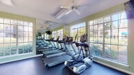 This fitness center at 1800 Grove Point Rd in Savannah, Georgia, is a bright, single-floor facility designed to support a comprehensive workout experience. The space is defined by its light sage-green walls, durable rubber flooring, and an abundance of natural light streaming through large multi-pane windows that frame views of lush surrounding greenery — creating an open, energizing atmosphere.

The cardio zone is well-appointed with multiple treadmills and elliptical machines arranged along a full-length mirrored wall, which amplifies the sense of space and allows users to monitor their form during workouts. Ceiling fans are positioned throughout to maintain comfortable airflow. Wall-mounted flat-screen televisions are conveniently placed for entertainment during cardio sessions.

The free weights area includes a full rack of dumbbells and kettlebells, complemented by a medicine ball and stability ball station, supporting a wide range of strength and functional training exercises. A multi-station cable and weight machine provides additional resistance training options. Motivational wall decals further enhance the energetic environment.

The facility also features a private interior door, an exit access point, and posted fitness center rules, reflecting a well-maintained and organized space. Overall, the fitness center offers a clean, inviting, and fully functional environment for residents to pursue their health and wellness goals.