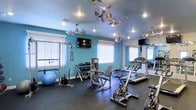 This fitness center is designed to support a comprehensive workout experience within a single, thoughtfully arranged floor. The room is finished with vibrant teal walls that lend an energetic ambiance, complemented by a dark rubber flooring ideal for high-impact exercise. Natural light filters in through multiple windows dressed with white blinds, brightening the space alongside recessed ceiling lights. A fleet of ceiling fans with metallic finishes ensures comfortable airflow throughout workouts. The equipment selection is well-rounded, featuring cardio machines including treadmills, a stationary bike, and a rowing-style cycle, alongside strength training options such as a cable pulley system, a leg press or extension machine, and a full dumbbell rack with a variety of weights. A stability ball and additional accessories round out the functional training options. Two wall-mounted flat-screen televisions are positioned for easy viewing from multiple workout stations, adding an entertainment element to the exercise environment. A large mirror on one wall expands the perceived depth of the room while allowing users to monitor their form. The overall layout is open and organized, making efficient use of the available space.