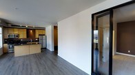 This single-floor space offers a clean, contemporary design with an efficient and open layout. The interior is anchored by rich, dark hardwood-style flooring that runs throughout, creating a cohesive and polished aesthetic.

The kitchen is positioned toward one end of the main living area and features warm honey-toned wood cabinetry, a stainless steel refrigerator, a dark countertop with an integrated sink, and track lighting overhead. The kitchen flows naturally into the open living area, making the space feel connected and functional.

A striking glass-paneled entry door with bold black framing serves as both a practical and design-forward feature, allowing natural light to filter into the interior while providing a glimpse of the outdoors. The crisp white walls throughout enhance the brightness and lend a fresh, modern feel to the space.

A hallway visible in the background suggests additional rooms or storage areas beyond the main living zone. The overall ambiance is clean and minimalist, with a balance of warm wood tones and modern industrial accents that give the space a versatile, move-in-ready character suitable for a variety of uses.