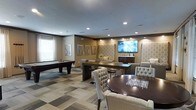 This well-appointed single-floor community lounge is designed to offer a refined and versatile gathering experience. The space is anchored by recessed lighting throughout the textured ceiling, casting a warm, inviting glow across the entire room. A patterned, multi-tone carpet adds visual interest underfoot and helps define the various functional zones within the space.

At the heart of the lounge is a dedicated recreation area featuring a full-size pool table, ideal for casual entertainment. Adjacent to it, a large flat-screen television is mounted on a feature wall accented with decorative wallpaper panels and warm ambient lighting, creating a natural focal point for social gatherings. A built-in credenza beneath the screen provides additional storage and display space.

The seating arrangement is equally thoughtful, with a mix of tufted upholstered chairs surrounding a dark wood round table in the foreground, and a longer rectangular dining-height table with bar-style seating positioned centrally. Additional lounge chairs are arranged near the TV wall, offering flexible configurations for both small and large groups. Natural light filters in through a window with horizontal blinds, complementing the overall warm, neutral color palette of tans, creams, and deep wood tones that give this amenity space a polished, welcoming character.