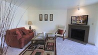 This single-floor residence offers a thoughtfully arranged living space designed for comfort and functionality. The living room serves as the heart of the home, featuring wall-to-wall carpeting that adds warmth underfoot and a classic fireplace that anchors the room as a natural focal point. Smooth, light-painted walls provide a clean, neutral backdrop that enhances the sense of openness throughout the space. The room comfortably accommodates a full seating arrangement, including a plush sofa and accent chair, centered around a glass-topped coffee table and layered with a geometric area rug that adds visual interest to the floor. Three framed wall art pieces are displayed symmetrically, contributing a curated, cohesive aesthetic to the space. Soft ambient lighting from a floor lamp and table lamp complements the overall warm tone of the room. The fireplace, finished with a simple mantel surround, provides both a functional heating element and a decorative centerpiece. The layout flows naturally, making efficient use of the available space while maintaining an uncluttered, welcoming atmosphere throughout the single level.