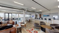 Located atop 1706 Washington Ave in St. Louis, MO, this rooftop clubhouse offers a sophisticated communal gathering space with an open, industrial-modern design. Exposed cylindrical ductwork runs overhead, complementing the warm hardwood-style flooring and bright white walls throughout the expansive main lounge. Large windows on multiple sides flood the interior with abundant natural light while framing panoramic views of the St. Louis skyline, including glimpses of the iconic Gateway Arch. The lounge area is furnished with a curated mix of leather sofas, velvet accent chairs, and patterned rugs arranged around a bold red accent wall with a wall-mounted television, creating an inviting social atmosphere. A dedicated dining area features a round wood table with contemporary seating, ideal for smaller gatherings. The full kitchen is outfitted with warm wood cabinetry, gray countertops, pendant lighting, stainless steel appliances, and a spacious white island with bar-height seating finished with gold-toned legs. Floor-to-ceiling sliding glass doors open directly onto a generous outdoor terrace, complete with a pergola structure, tropical plantings, and alfresco dining and lounge seating — offering unobstructed views of the surrounding cityscape. St. Louis-themed artwork is displayed throughout, reinforcing a strong sense of local identity.