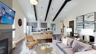 This single-floor space showcases a thoughtfully designed open-concept layout that seamlessly integrates the living and kitchen areas into one cohesive, flowing environment. The most striking architectural feature is the vaulted ceiling adorned with dark-stained exposed wooden beams, which add a dramatic contrast against the crisp white ceiling and walls above.

The kitchen is well-appointed with dark cabinetry, stainless steel appliances including a French door refrigerator and microwave, and a statement range hood in white. A large island with a light wood panel finish and bar-height seating serves as both a functional workspace and a natural dividing point between the kitchen and living areas. A series of pendant lights hang at varying heights above, complementing the recessed lighting throughout.

The living area features rich dark hardwood flooring, an area rug, and carefully chosen furnishings including a grey sofa and a warm cognac leather accent chair. Natural light filters in through windows along the perimeter, enhancing the bright, airy ambiance. The overall aesthetic balances modern sophistication with warm, inviting elements.