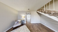 This two-story home offers a well-organized layout across two floors, with the main living areas on the ground level and private bedrooms above. The lower floor features an open-concept kitchen and living room finished with rich wood-look flooring throughout. The living room is bright and welcoming, highlighted by a large front-facing window that invites natural light and offers outdoor views. A staircase with white railings connects the two levels seamlessly. The kitchen is equipped with white cabinetry, dark granite-style countertops, black appliances including a range and dishwasher, and an under-cabinet range hood — a functional and clean workspace. One full bathroom on the main floor features a tub-shower combo with tile surround and a separate vanity area with a white cabinet and large wall mirror. Upstairs, two carpeted bedrooms provide comfortable retreats. The primary bedroom is generously sized and features floor-to-ceiling mirrored sliding closet doors and a ceiling fan. The second bedroom is bright and versatile with wood-look flooring. A hallway connects the upper-level rooms, with a second full bathroom nearby. Textured ceilings and neutral wall tones are consistent throughout, creating a cohesive and practical living environment.