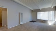 This single-floor space showcases a distinctive architectural character defined by its vaulted ceiling with exposed white-painted wooden beams and tongue-and-groove paneling, lending a classic mid-century aesthetic. The living area is finished with wall-to-wall carpeting in a neutral tone, providing a warm and comfortable underfoot feel. A wall-mounted heater unit is positioned along one wall, serving as the primary heating source for the space. The interior is painted in a soft, neutral gray, creating a clean and versatile backdrop throughout. Natural light filters generously into the space through a large window fitted with vertical blinds near the entryway, brightening the interior while offering privacy control. The front entry door is centrally visible from the main living area, contributing to a straightforward and open floor plan. The overall ambiance is simple, functional, and well-lit, with the exposed beam ceiling serving as the standout architectural detail that adds height and visual interest to the living space.