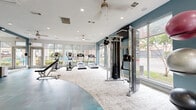 This modern fitness center offers a thoughtfully designed workout environment spread across a single open floor. The space is defined by a striking dual-tone flooring scheme, combining teal-hued resilient flooring in the free weights and functional training zones with a light, textured carpet in the cardio and stretching areas. Large floor-to-ceiling windows wrap around multiple walls, flooding the interior with abundant natural light and providing views of the surrounding greenery and outdoor areas.

The cardio section is well-appointed with multiple treadmills and elliptical trainers positioned along the windowed perimeter, allowing users to enjoy outdoor views during their workouts. Ceiling fans are mounted overhead, complementing the recessed lighting for a comfortable exercise environment. A cable machine and functional trainer station occupy a central area, supported by an adjustable weight bench nearby.

The free weights zone features a fully stocked dumbbell rack alongside kettlebells and a medicine ball storage rack. An athletic-themed black-and-white mural adds an energetic, motivational aesthetic to one wall, while a wall-mounted television provides entertainment. Full-length mirrors reflect light and enhance the sense of spaciousness. The overall ambiance is clean, bright, and purpose-built for a comprehensive fitness experience.