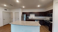This single-floor unit offers a well-designed open layout with consistent finishes throughout. The kitchen serves as a central highlight, featuring rich espresso-toned cabinetry extending to the upper walls, light granite countertops with subtle veining, and a suite of stainless steel appliances including a refrigerator, dishwasher, microwave, and range. A large granite-topped island provides additional prep and seating space, while recessed lighting illuminates the area effectively. Wood-style flooring runs through the kitchen and connects seamlessly to the open living area, which benefits from a bank of windows fitted with horizontal blinds, allowing generous natural light to fill the space. A French door adds an architectural detail, enhancing the flow between areas. The bedroom features plush carpeting, a ceiling fan with light fixture, and a window with an outdoor view. An additional room with wood-style flooring and a single window offers flexible use. The bathroom is finished with matching espresso cabinetry, a granite-topped vanity, a large framed mirror, and a full tub-shower combination, maintaining a cohesive aesthetic consistent with the rest of the unit.
