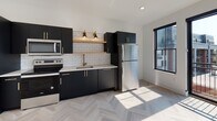 This contemporary single-floor unit showcases a clean, modern aesthetic throughout, highlighted by light-toned herringbone hardwood flooring that flows seamlessly across the living areas and bedroom. The open living space is bathed in natural light from large black-framed sliding glass doors that lead to a private balcony, offering open rooftop views of the surrounding neighborhood. A sleek ceiling fan adds both function and style overhead.

The kitchen is a standout feature, designed with matte black cabinetry accented by gold hardware, an open wood floating shelf, classic white subway tile backsplash, and a gold-toned faucet. Stainless steel appliances — including a refrigerator, range, and microwave — complete the space, which also benefits from natural light through a large black-framed window.

The bedroom offers generous closet storage with a single door and a set of double doors, complemented by recessed lighting and the same elegant herringbone flooring.

The bathroom is finished with large-format white hexagonal floor tiles, a matte black vanity with gold hardware, a matching black-framed mirror, and a warm wall sconce, creating a refined monochromatic contrast. Throughout the unit, the consistent use of black, white, and gold tones delivers a cohesive, upscale modern design.