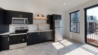 This contemporary single-floor unit showcases a clean, modern aesthetic throughout, highlighted by light-toned herringbone hardwood flooring that flows seamlessly across the living areas and bedroom. The open living space is bathed in natural light from large black-framed sliding glass doors that lead to a private balcony, offering open rooftop views of the surrounding neighborhood. A sleek ceiling fan adds both function and style overhead.

The kitchen is a standout feature, designed with matte black cabinetry accented by gold hardware, an open wood floating shelf, classic white subway tile backsplash, and a gold-toned faucet. Stainless steel appliances — including a refrigerator, range, and microwave — complete the space, which also benefits from natural light through a large black-framed window.

The bedroom offers generous closet storage with a single door and a set of double doors, complemented by recessed lighting and the same elegant herringbone flooring.

The bathroom is finished with large-format white hexagonal floor tiles, a matte black vanity with gold hardware, a matching black-framed mirror, and a warm wall sconce, creating a refined monochromatic contrast. Throughout the unit, the consistent use of black, white, and gold tones delivers a cohesive, upscale modern design.