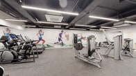 This fitness center offers a generously sized, single-floor workout space designed to accommodate a comprehensive range of exercise routines. The open floor plan is finished with durable rubber flooring throughout, providing a safe and functional surface for all types of training activities.

The space is outfitted with an extensive selection of equipment, including selectorized strength machines, cable systems, leg press units, adjustable benches, stationary bikes, treadmills with integrated screens, and rowing machines. A dedicated functional training area features stability balls, medicine balls, and foam rollers stored on a centrally positioned rack, offering versatility for group or individual workouts.

Large wall-mounted mirrors span multiple walls, enhancing the sense of spaciousness and allowing users to monitor their form during exercise. The ceiling features an exposed industrial design with dark-painted ductwork and sleek linear LED lighting that ensures bright, even illumination across the entire space. A cassette-style HVAC unit provides climate control overhead.

A striking full-wall mural depicting dynamic athletic figures in motion adds an energetic and motivating visual element to the space. The overall design is clean, modern, and purposefully functional, creating an environment well-suited for focused, high-quality training.