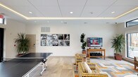 This single-floor community space is thoughtfully designed to serve as a multifunctional gathering hub, combining recreation, dining, and relaxation within a bright, contemporary setting. The layout flows seamlessly across light-toned tile and wood-style flooring, creating a cohesive and inviting atmosphere throughout.

The lounge area anchors the space with a collection of colorful, patterned armchairs and a leather sofa arranged around a geometric area rug in warm orange and grey tones. A wall-mounted flat-screen television is positioned above a wooden media console, flanked by tall indoor plants that add a natural element. A curated grid of black-and-white photography and vibrant abstract artwork decorates the walls, lending the space a distinct artistic character.

Large windows and clerestory windows line multiple walls, flooding the interior with abundant natural light while offering views of an outdoor pool area and surrounding streetscape. Recessed lighting and subtle LED cove lighting along the ceiling tray provide warm, layered illumination.

A ping pong table occupies a dedicated game zone adjacent to the lounge, accompanied by high-top bistro seating for casual dining or socializing. The kitchen features warm wood cabinetry, dark tile backsplash, stainless steel appliances, pendant lighting, and a spacious island with bar stool seating, making it well-suited for group gatherings and events.