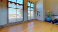 This yoga and fitness room is a thoughtfully designed studio space ideal for yoga, stretching, and group exercise. The room immediately impresses with its soaring ceilings that create an open, airy atmosphere, while the warm orange accent wall adds an energizing pop of color that complements the neutral tones throughout. Underfoot, smooth hardwood flooring provides a clean, durable surface well-suited for a variety of fitness activities. A large wall of floor-to-ceiling mirrors spans one side of the room, allowing practitioners to monitor form and technique while also visually expanding the space. Abundant natural light pours in through oversized, multi-pane windows that reach toward the ceiling, offering views of the outdoors and contributing to the bright, welcoming ambiance. The studio is equipped with a rack of exercise and stability balls in an array of sizes, supporting diverse workout routines beyond yoga alone. Framed artwork adorns the walls, adding a refined touch to the functional environment. A set of double glass doors provides easy access, seamlessly connecting the studio to adjacent areas. Overall, this yoga room strikes a balance between a calm, focused atmosphere and an energizing setting designed to inspire movement and wellness.