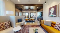 Located at 305 Prine Pl in Charlotte, NC, this single-floor amenity space is a thoughtfully designed communal hub that blends modern aesthetics with practical functionality. The open-plan layout is anchored by a central lounge area featuring a bold mustard leather sofa, high-back privacy seating pods, and layered plush area rugs atop polished concrete floors—creating distinct zones for both relaxed gathering and focused work.

Rich wood-paneled walls and ceilings lend warmth and a refined, contemporary character throughout the space, complemented by recessed and pendant lighting that sets a welcoming ambiance. A large wall-mounted display screen serves as a focal point in the main lounge, while curated abstract artwork adds a gallery-like quality to the bright white accent walls.

A private conference room, enclosed with sleek glass-paneled doors, features a long dark conference table lined with mustard upholstered chairs, wood accent walls, linear ceiling lighting, and its own wall-mounted display—ideal for meetings and presentations. Sheer floor-to-ceiling curtains along the windowed perimeter diffuse abundant natural light throughout the space. Lush indoor plants are positioned throughout, adding life and a touch of nature to this polished, design-forward environment.