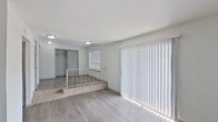 This single-floor space features a thoughtfully designed split-level layout, where a raised platform area with a metal safety railing and stone-faced steps creates a distinct visual separation between living zones. Light-toned wood-style flooring runs throughout, lending a clean, cohesive look to the interior.

The main living area is bright and open, benefiting from a large sliding glass door dressed with vertical blinds that allows generous natural light to fill the space. An additional wide window in the lower level further enhances the sense of openness and provides views to the exterior.

A pass-through window connects the kitchen to the living area, offering a convenient and functional layout. The kitchen is positioned along one wall with cabinetry and appliances visible through the opening.

A built-in closet with rod and shelf storage is accessible from the main living space. The bathroom features wood-style flooring, a white vanity with under-sink cabinetry, a wall-mounted mirror, and a lighted fixture above, offering a clean and functional design. Flush-mount ceiling lights throughout provide consistent, practical illumination across all areas of the space.