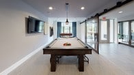 This single-floor game room is thoughtfully designed as a dedicated recreational and social space. At its center sits a full-size pool table with a neutral felt surface, complemented by a wall-mounted cue rack and a scoring tally sheet nearby. A wall-mounted flat-screen television provides additional entertainment options along one side of the room.

Toward the windowed end of the space, a foosball table is positioned beneath a large window dressed with steel-blue curtains, allowing natural light to fill the area and offering views of the outdoors. The room is accessed via a set of sleek sliding barn-style glass doors, lending a contemporary industrial touch to the otherwise clean, modern aesthetic.

A cozy lounge nook is tucked into one corner, featuring a cushioned banquette, round bistro-style tables, and wooden chairs — ideal for casual seating between games. A bold black-and-white artwork featuring Albert Einstein and the phrase "Games Make You Smart" anchors this corner with personality and playfulness.

The space is finished with neutral carpeted flooring, white walls, and recessed lighting throughout, creating a bright and inviting atmosphere. The overall design balances functionality with a modern, social ambiance well-suited for group recreation and leisure.