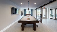 This single-floor game room is thoughtfully designed as a dedicated recreational and social space. At its center sits a full-size pool table with a neutral felt surface, complemented by a wall-mounted cue rack and a scoring tally sheet nearby. A wall-mounted flat-screen television provides additional entertainment options along one side of the room.

Toward the windowed end of the space, a foosball table is positioned beneath a large window dressed with steel-blue curtains, allowing natural light to fill the area and offering views of the outdoors. The room is accessed via a set of sleek sliding barn-style glass doors, lending a contemporary industrial touch to the otherwise clean, modern aesthetic.

A cozy lounge nook is tucked into one corner, featuring a cushioned banquette, round bistro-style tables, and wooden chairs — ideal for casual seating between games. A bold black-and-white artwork featuring Albert Einstein and the phrase "Games Make You Smart" anchors this corner with personality and playfulness.

The space is finished with neutral carpeted flooring, white walls, and recessed lighting throughout, creating a bright and inviting atmosphere. The overall design balances functionality with a modern, social ambiance well-suited for group recreation and leisure.